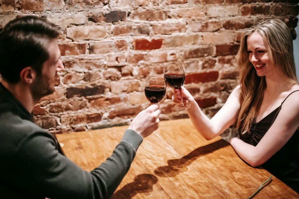 Young couple in elegant clothes sitting at table in restaurant near brick wall and enjoying date while proposing toast with r