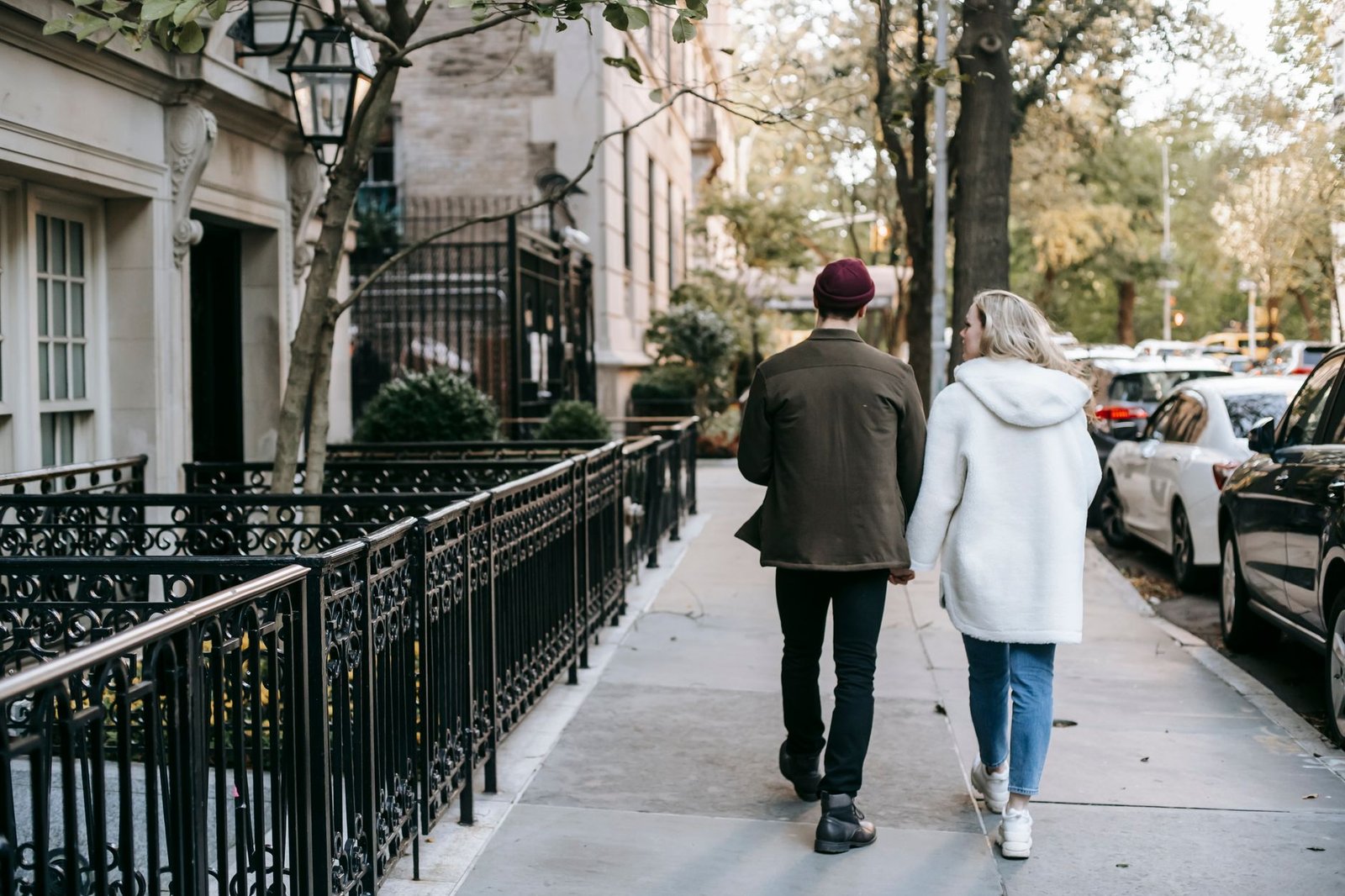Back view of anonymous trendy young couple in stylish warm clothes holding hands and walking in city street near residential