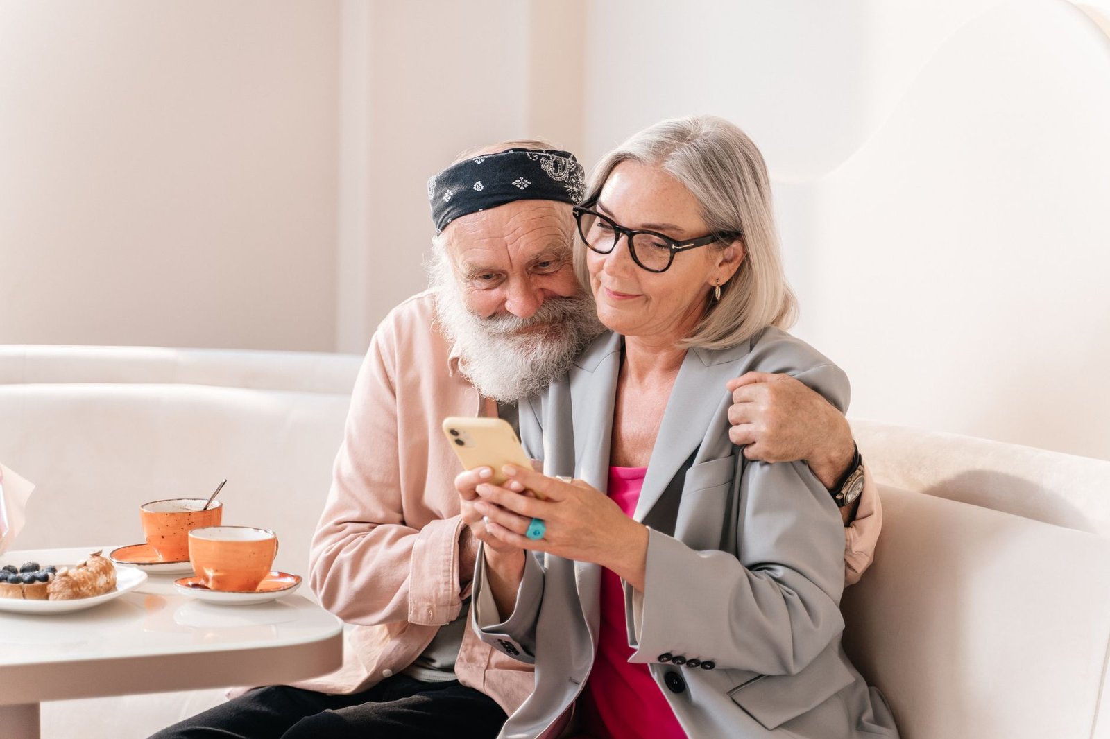 Senior couple embracing and smiling while using a smartphone in a cozy cafe setting.