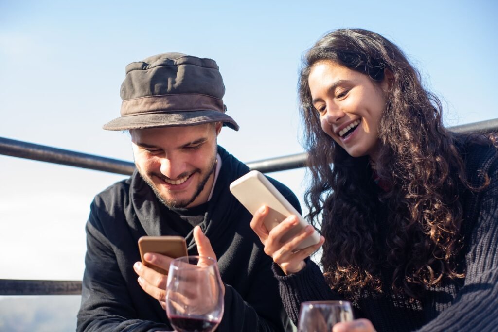 Two friends smiling and using smartphones while enjoying a drink outdoors, capturing a joyful moment.