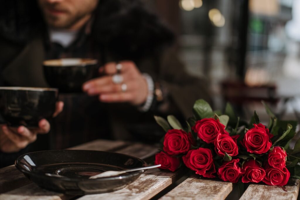 Crop unrecognizable man and woman in stylish outfit drinking coffee in street cafe sitting at table with bunch of elegant red