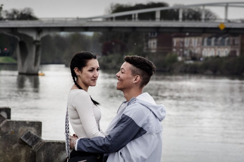 Young couple sharing a tender moment by a scenic riverside with a bridge in the background.