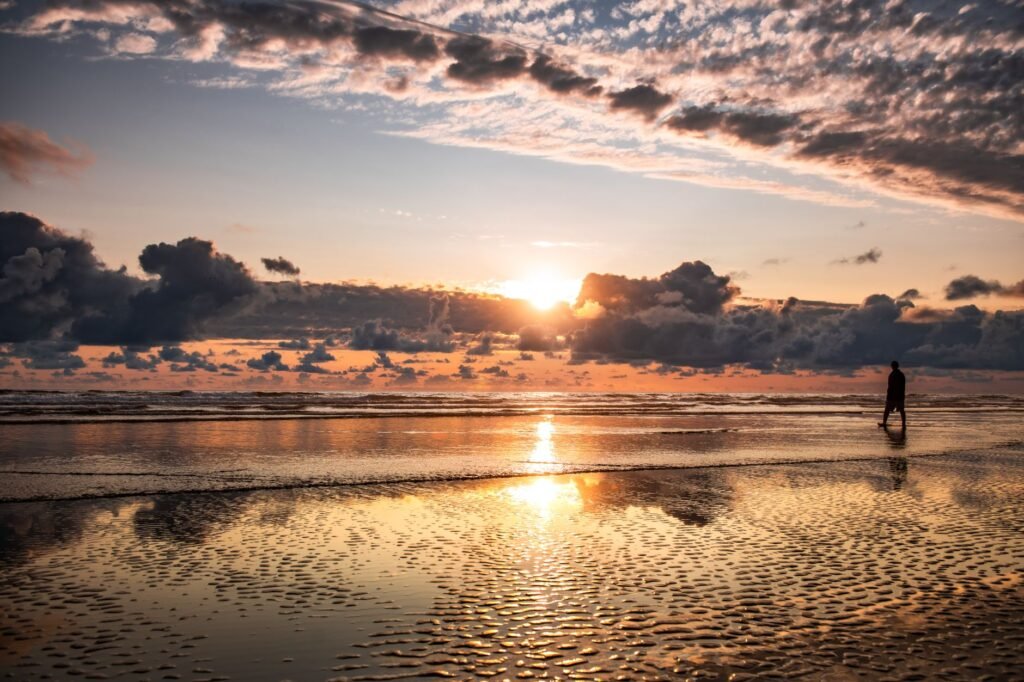 Silhouette of a person walking along Cannon Beach with a stunning sunset and dramatic clouds reflecting on wet sand.