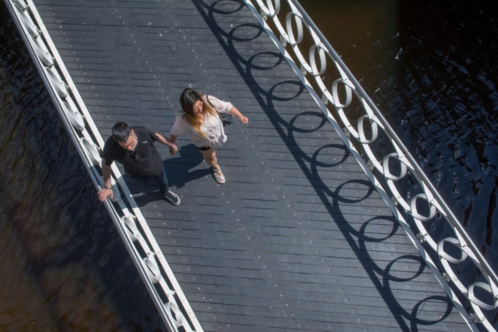 A couple holds hands and walks across a modern footbridge on a sunny day.