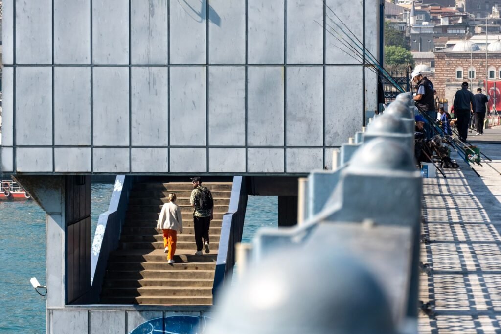 People walking on bridge stairs with waterfront views and fishing activities.