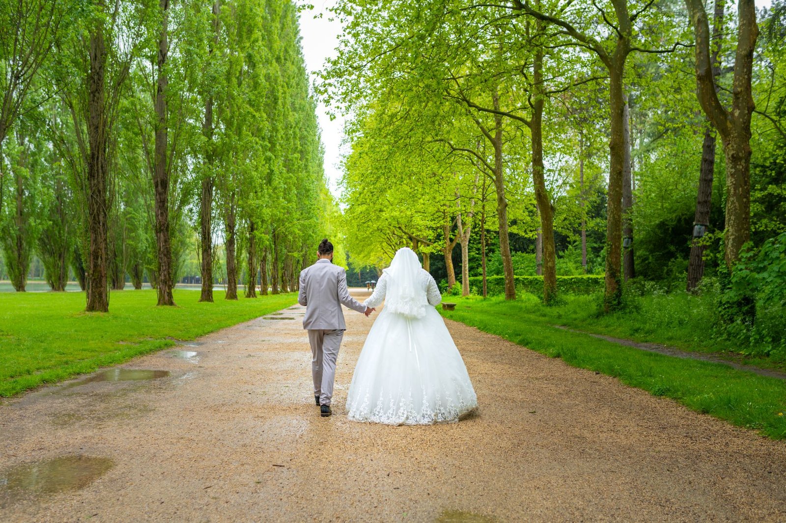 Bride and groom walking hand in hand down a tree-lined path in a lush green park, symbolizing love and togetherness.