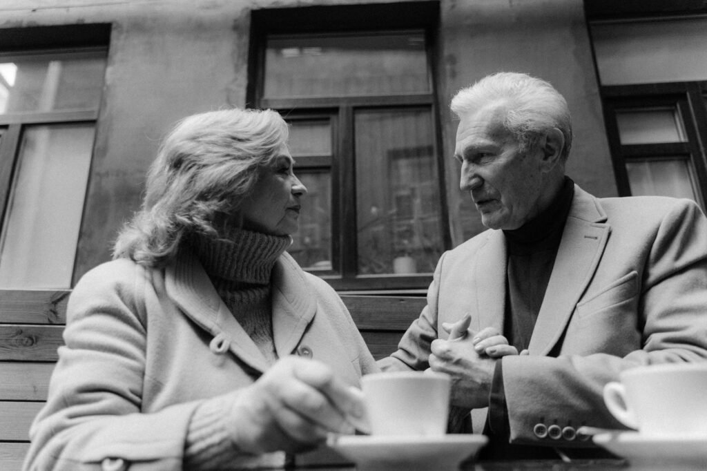 A black and white photo of a senior couple holding hands while having coffee outdoors.