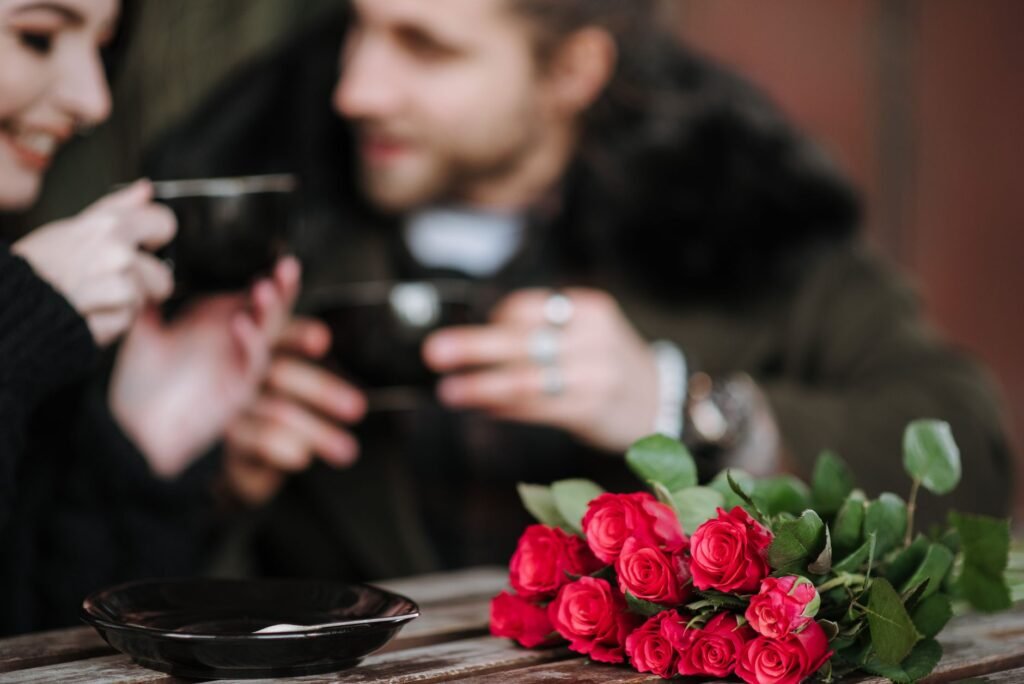 Crop man talking to smiling girlfriend with hot drink at cafeteria table with blooming rose bouquet