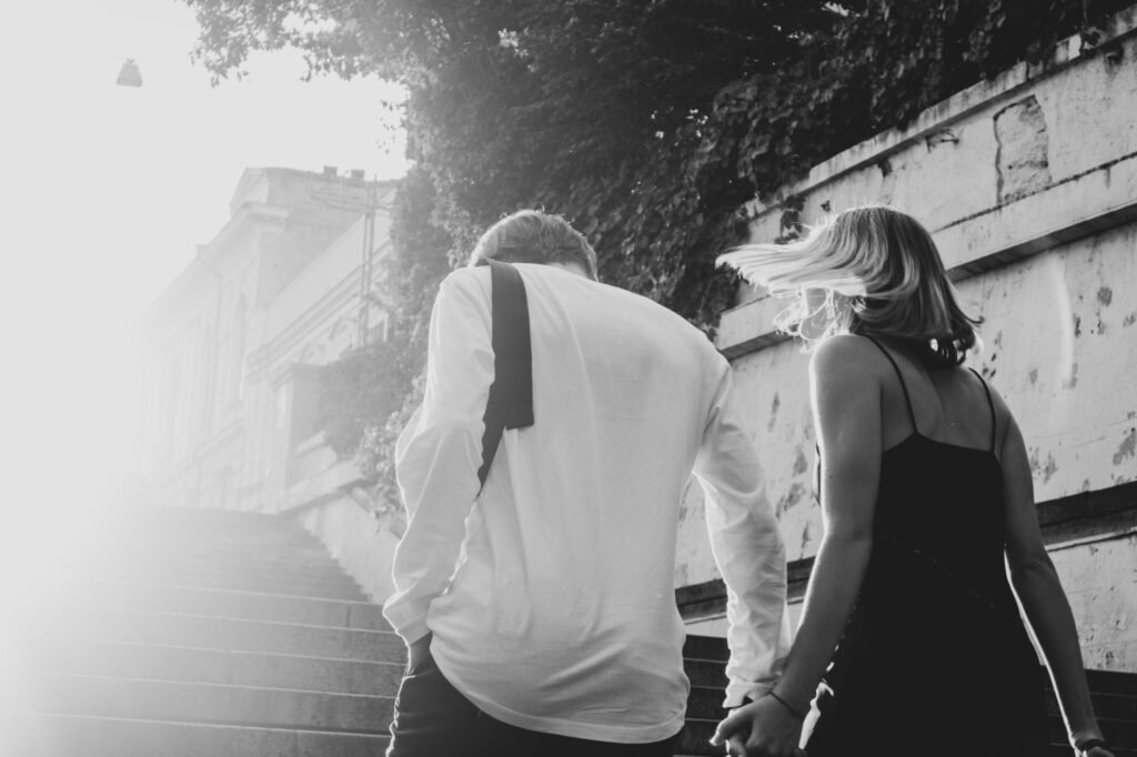 Couple holding hands, walking up sunlit steps, capturing romance in a monochrome setting.