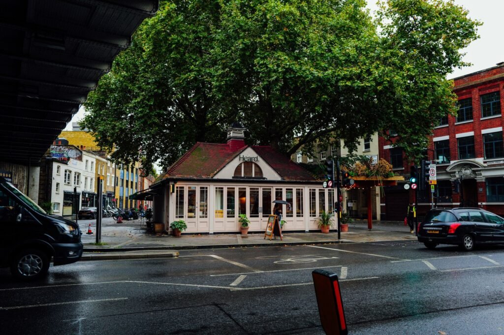 A quaint cafe nestles under a large tree on a rainy street corner, offering a cozy urban retreat.