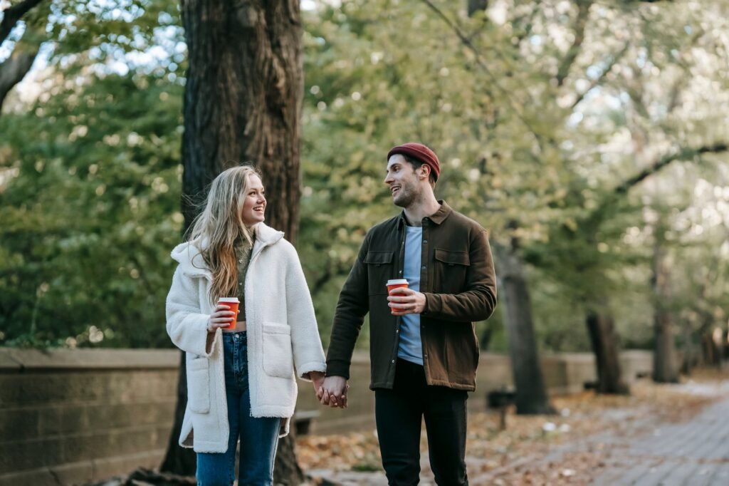 Happy stylish young man and woman in warm clothes holding hands and chatting while walking in autumn park and drinking takeaw