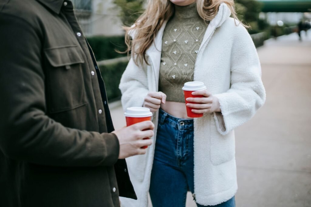 Crop faceless young female and male in stylish outfits standing on city street and drinking coffee to go during break