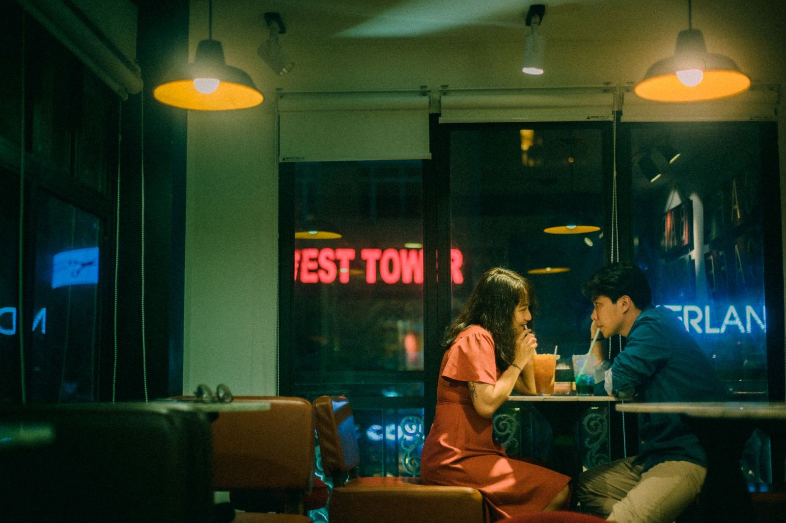 Side view of smiling ethnic woman and boyfriend drinking refreshing beverages through straws while sitting at cafeteria table