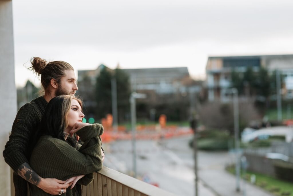 Side view of young unshaven male embracing female partner while admiring city from fenced balcony and looking away