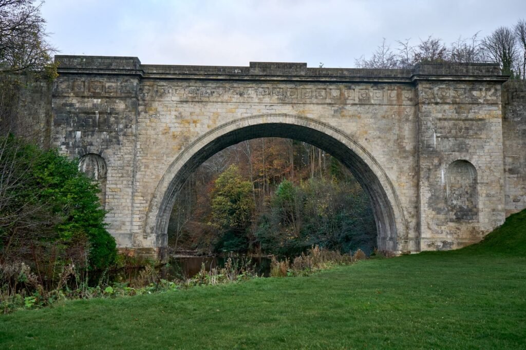 A historic stone arch bridge set in a scenic rural park with lush greenery and an overcast sky.