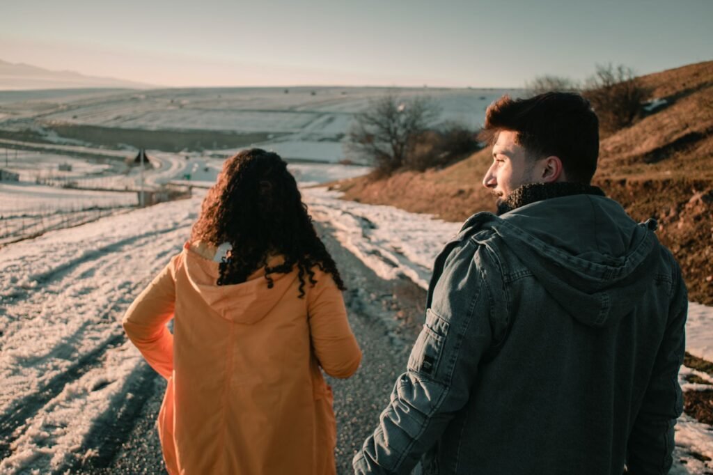 Back view of young couple of travelers in warm outerwear walking along path in mountainous valley covered with snow
