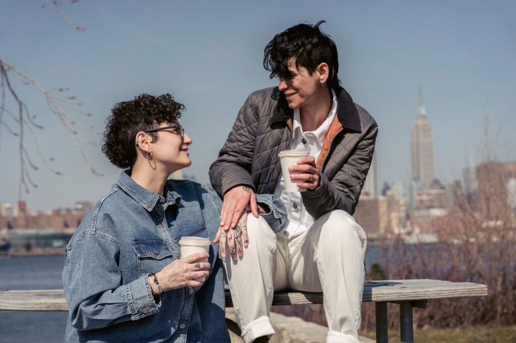 Enamored young lesbian couple with short dark hair in trendy outfits smiling and looking at each other while resting on woode