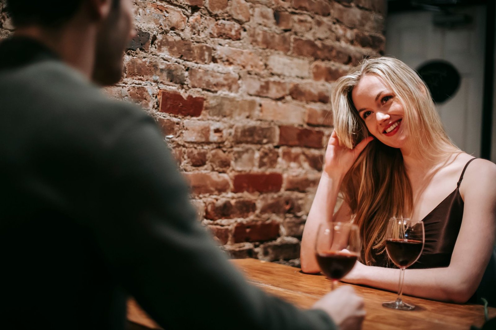 Young couple in elegant outfits sitting in light cafeteria at wooden table near brick wall and having romantic dinner with re