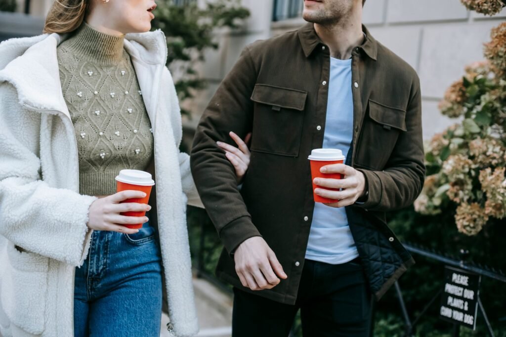 Crop anonymous young couple in stylish clothes drinking takeaway coffee and talking while walking together in city street dur