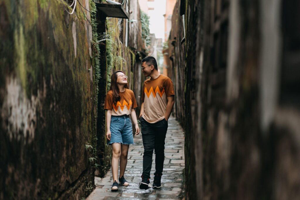 Full body of positive Asian couple in casual same outfit and spectacles walking on narrow street near shabby walls and lookin