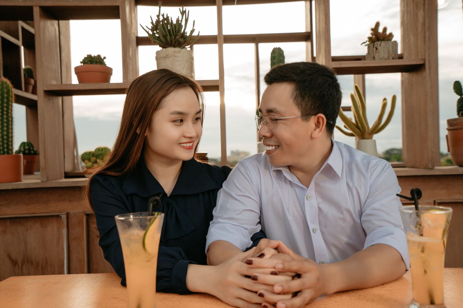 Smiling Asian couple in elegant clothes sitting at table with drinks in cafeteria and holding hands while looking at each oth