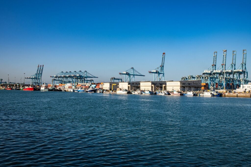 A bustling harbor with large cranes, cargo containers, and ships under a clear blue sky.