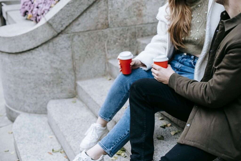 Unrecognizable young couple sitting on steps outside building and drinking hot beverage in paper cups