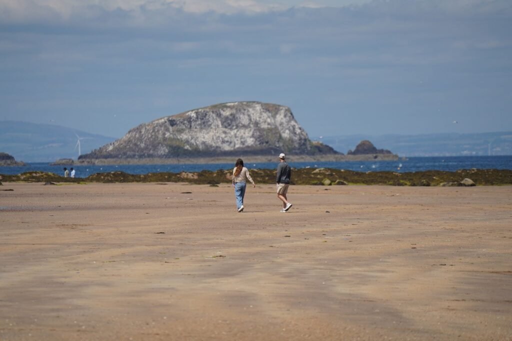 Couple strolling on a sandy beach with Bass Rock in the background on a sunny day in North Berwick, Scotland.