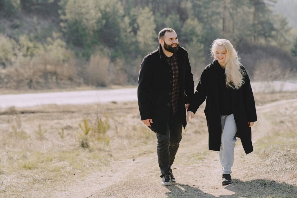 Full body cheerful couple in warm clothes holding hands and smiling while strolling together in countryside during romantic d