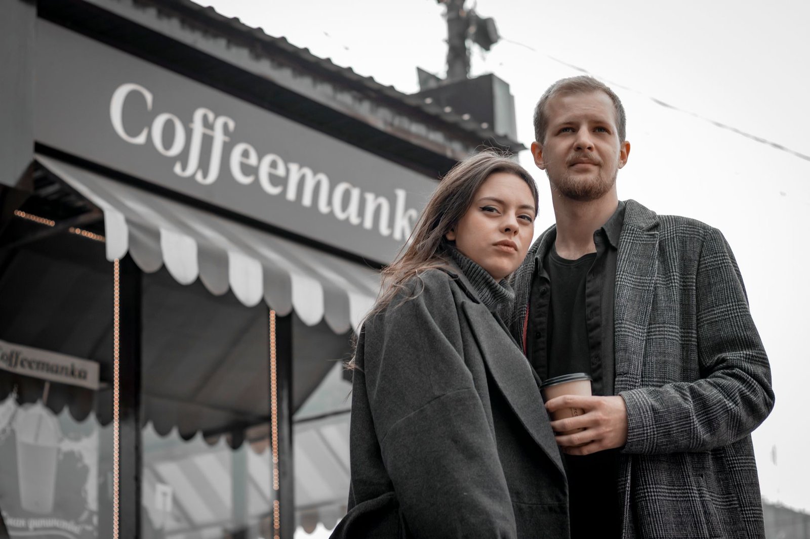 Young couple in coats standing outside Coffeemanka café, holding coffee, in an urban setting.