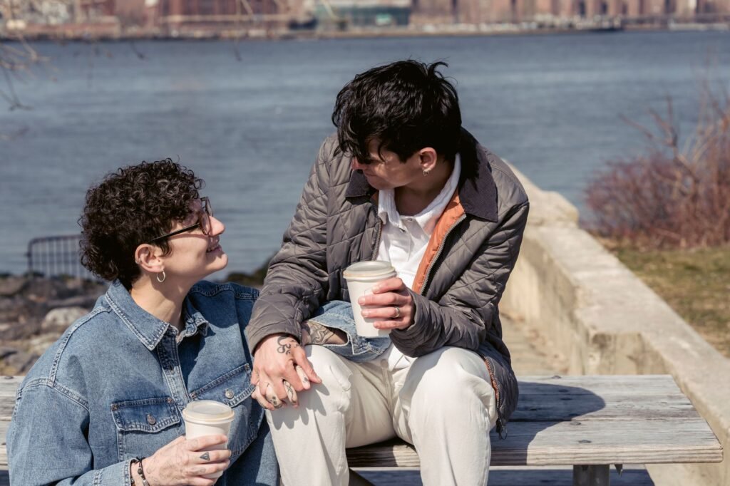 Enamored young smiling lesbian couple with short dark hair holding hands and looking at each other while drinking takeaway co