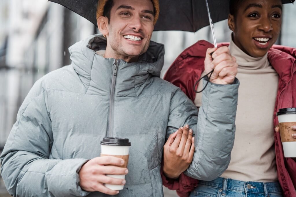 Crop multiethnic positive couple with carton cups of coffee smiling and promenading under umbrella while looking away on blur