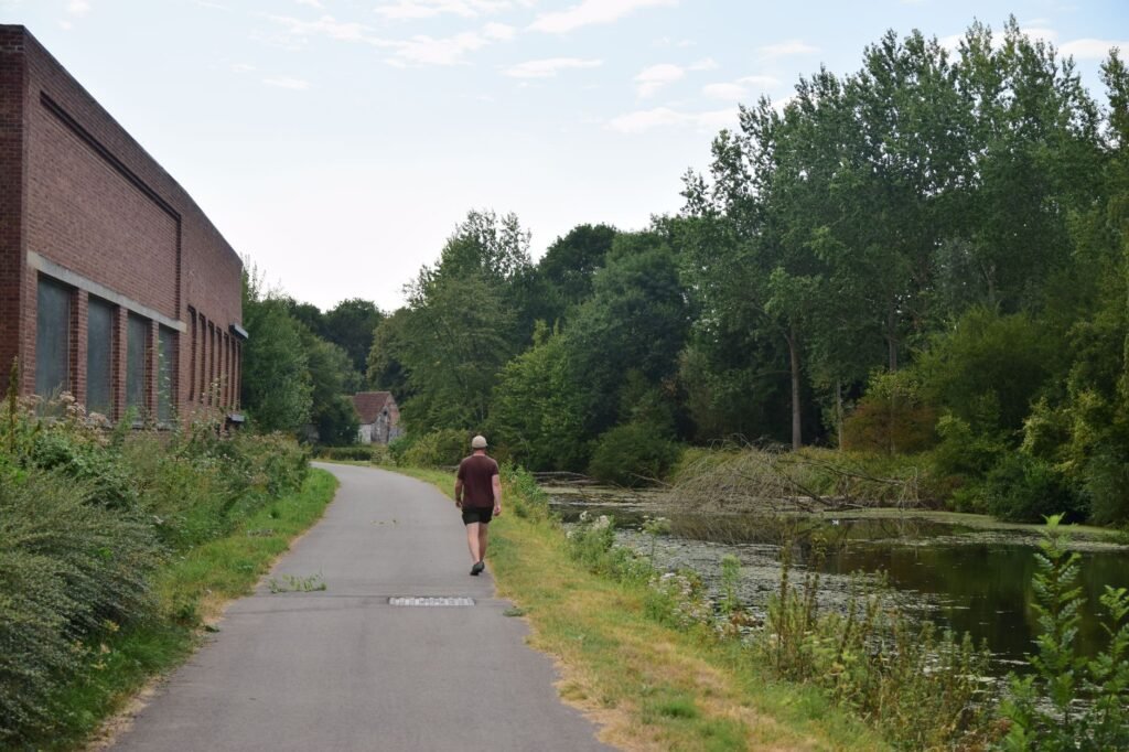 A person walking on a riverside path next to a red brick building and lush greenery.