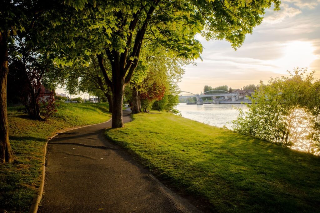 Beautiful sunset view of a riverside park with trees, a path, and a bridge in Compiègne, France.