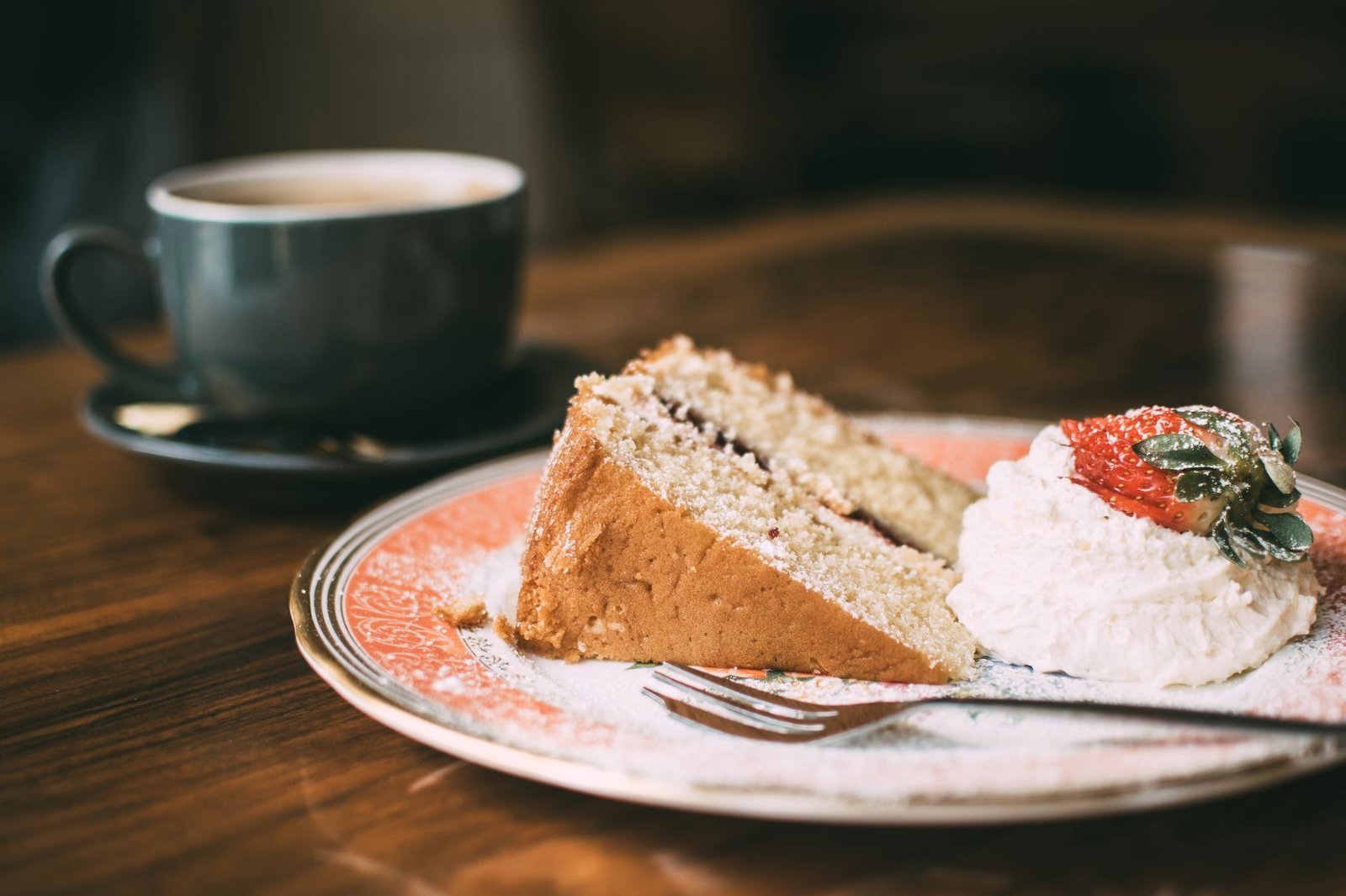 A slice of sponge cake with cream and strawberry, served with a hot cup of coffee, in a cozy café setting.