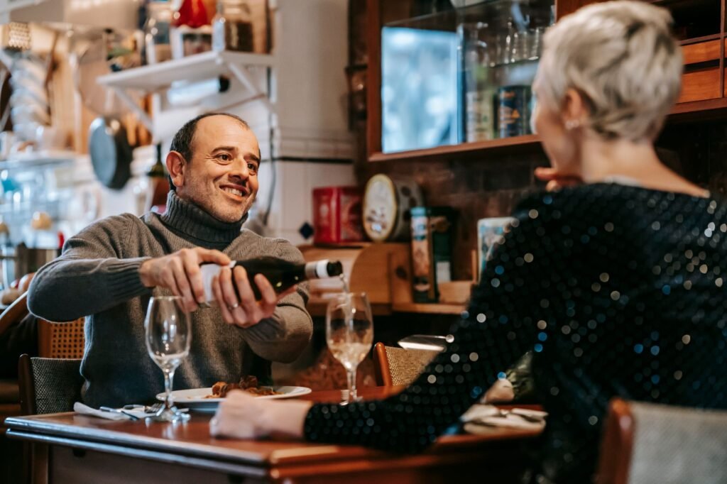 Positive couple having date at table while man pouring wine from bottle into wineglasses near plates with tasty dish in brigh