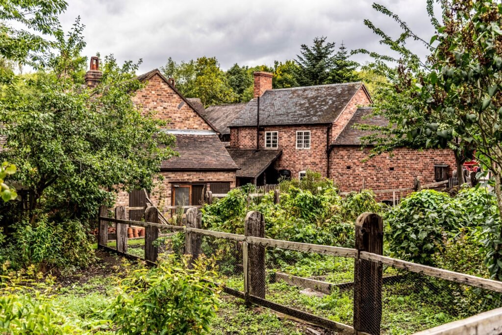Quaint English brick cottage surrounded by a lush green garden with wooden fencing.