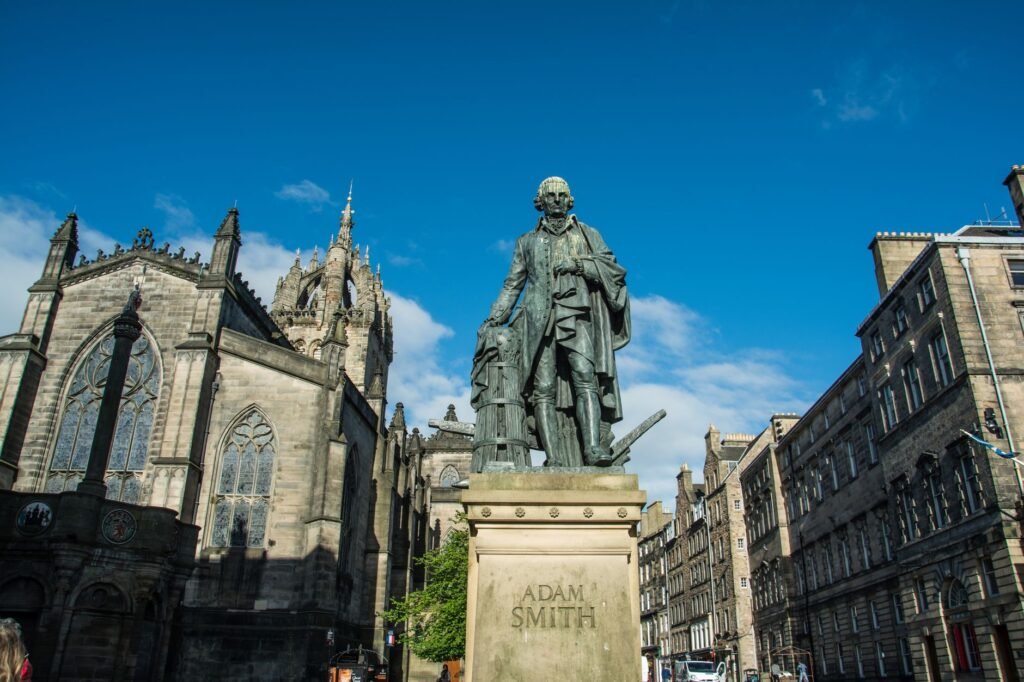Statue of Adam Smith in Edinburgh city center with historical architecture and vibrant blue sky.
