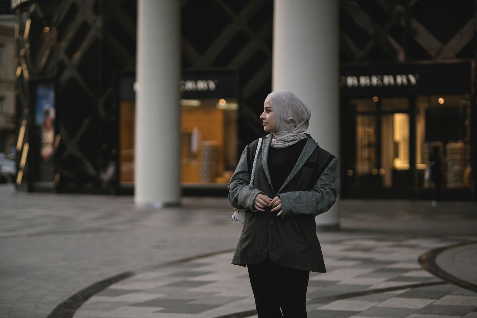Stylish woman wearing hijab poses outside a modern shopping mall in the city.