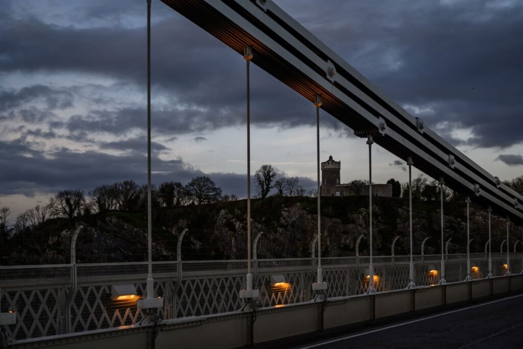 A scenic twilight view of a historic suspension bridge in the UK with a castle in the background.