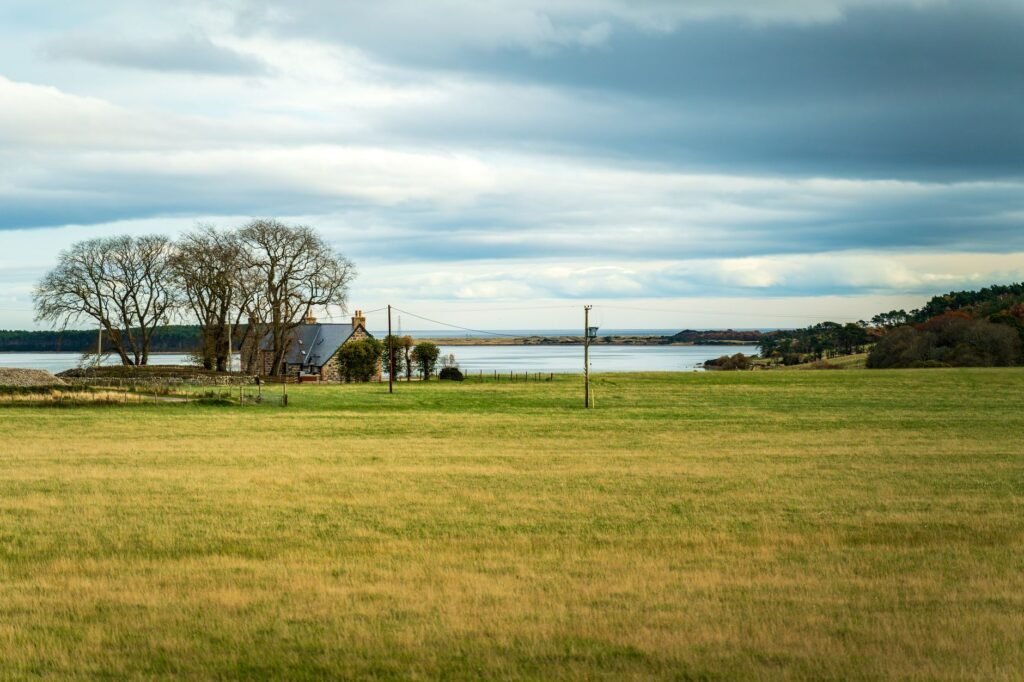 Peaceful rural scene featuring a farmhouse by a serene lake with open fields and cloudy skies.