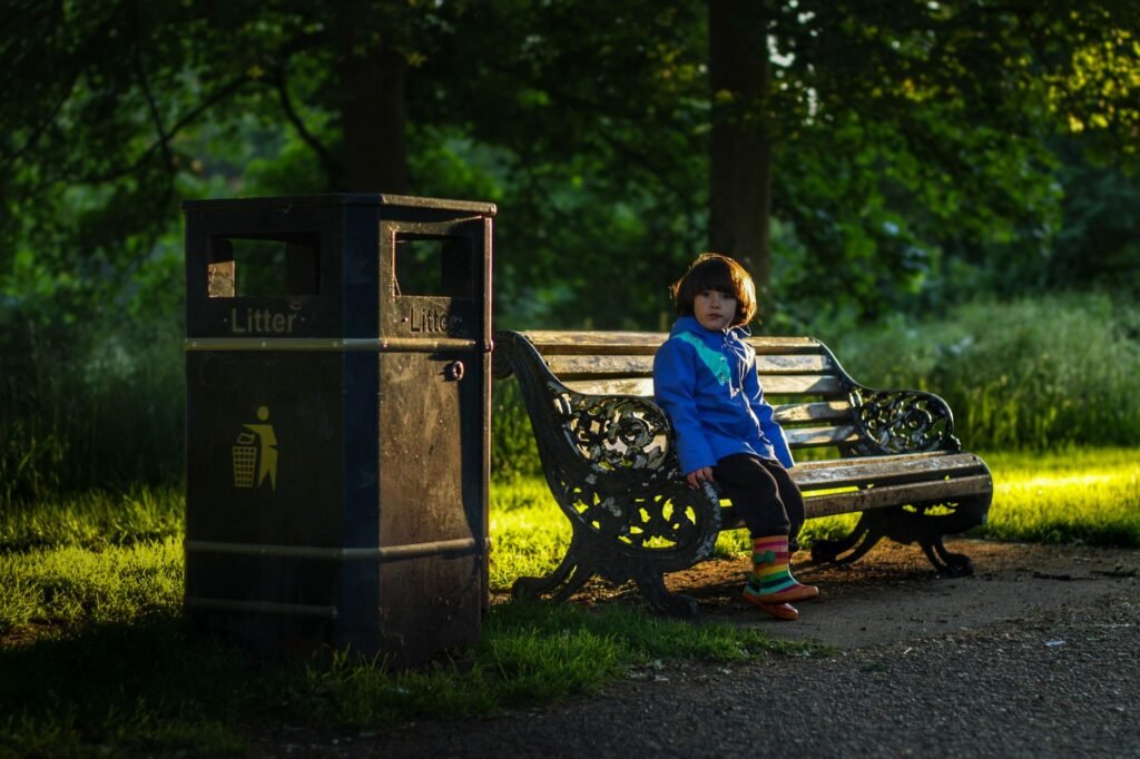 A child in a blue jacket sits on a park bench beside a litter bin under warm evening light.