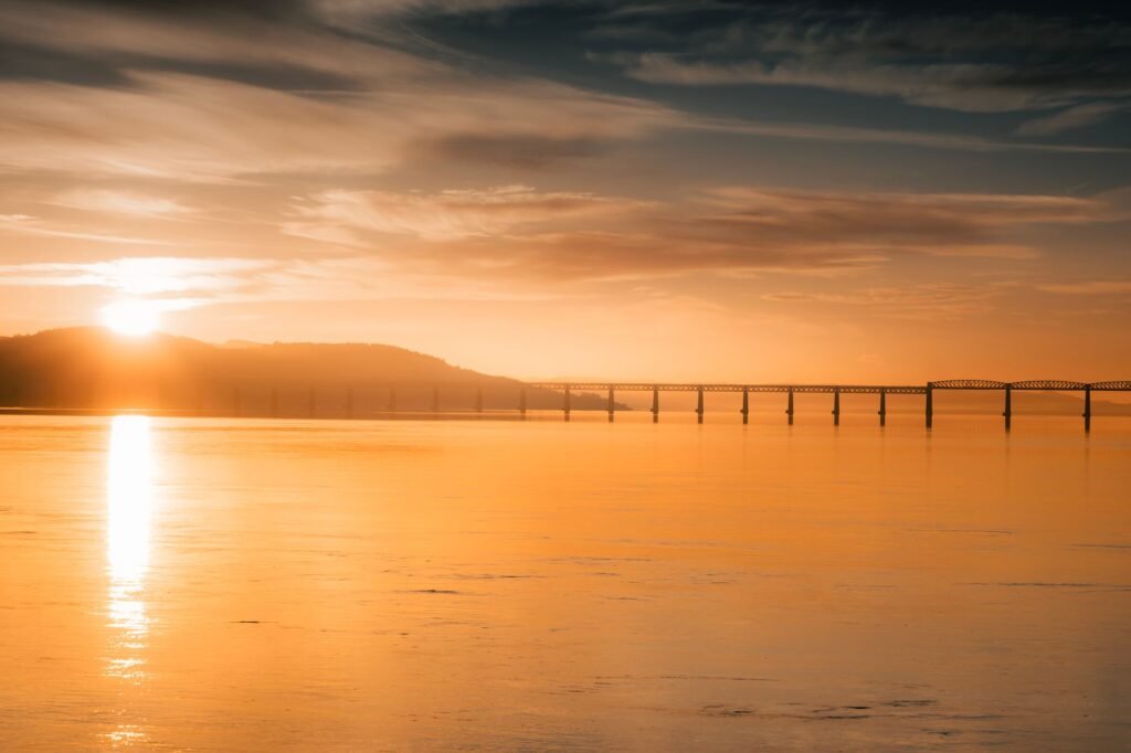 Golden sunset view of Forth Bridge reflecting on calm waters in Scotland, capturing natural beauty.
