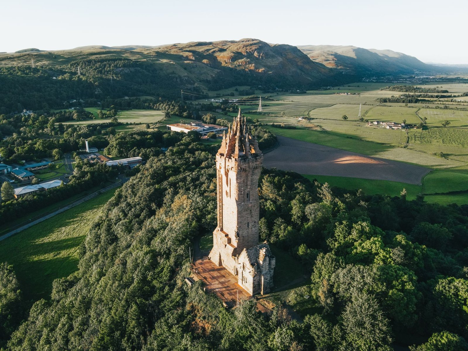 Aerial shot of the historic Wallace Monument surrounded by lush greenery in Scotland, captured at golden hour.