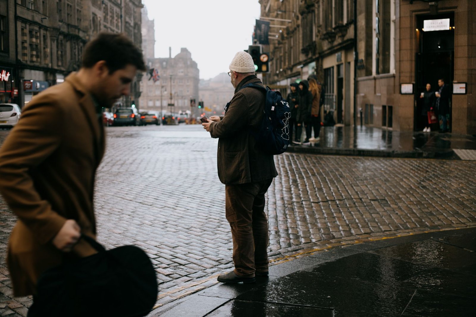 Young pedestrian walking on cobblestone roadway near unrecognizable male and aged buildings on rainy day
