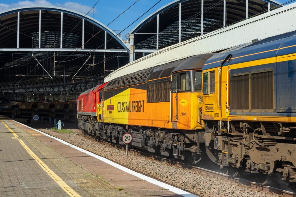 Freight trains with vibrant liveries at a railway station platform under a sunny sky.