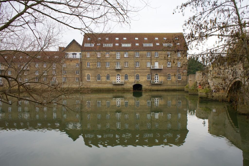 A tranquil view of a historic building reflecting on a serene river in Godmanchester, England.