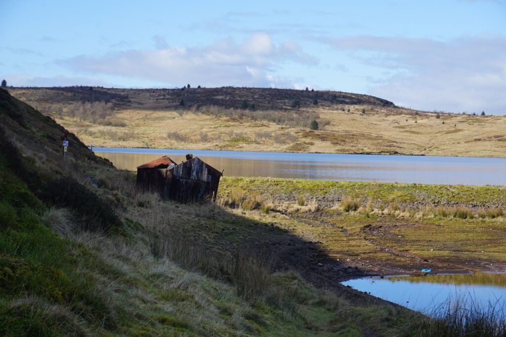 Rustic shack by a tranquil lake in the Scottish Highlands with scenic hills in the background.