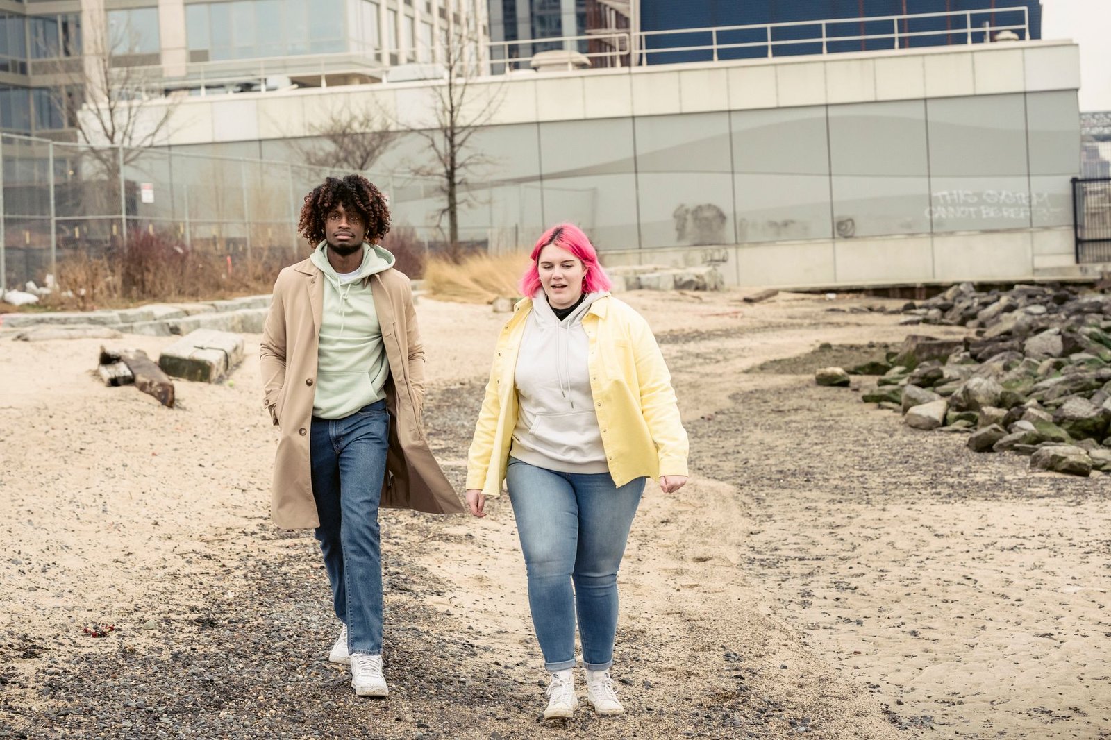 Full body of multiracial couple in jeans and outerwear strolling on embankment with stones against building during date in ci