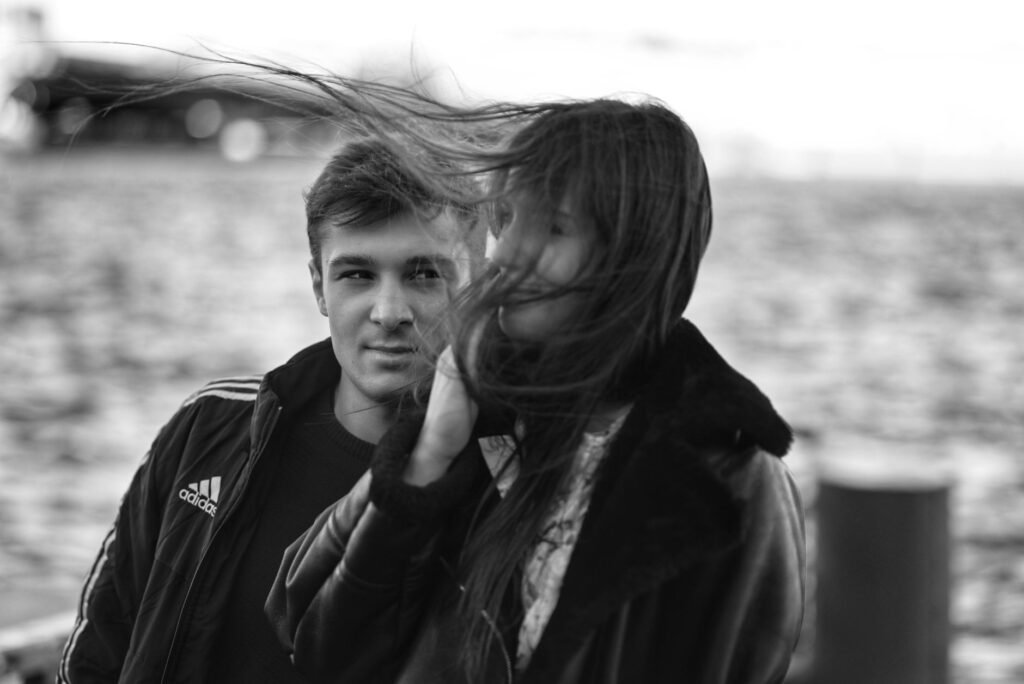 Moody black and white portrait of a couple standing outdoors, hair blowing in the wind.